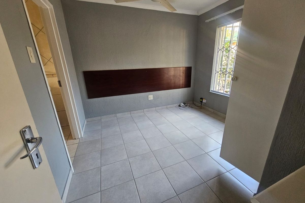 A clean, empty bedroom featuring tiled flooring, a ceiling fan, and a built-in headboard, with an adjacent doorway leading to a bathroom.