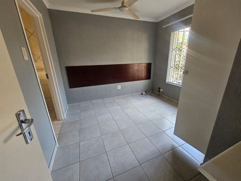 A clean, empty bedroom featuring tiled flooring, a ceiling fan, and a built-in headboard, with an adjacent doorway leading to a bathroom.