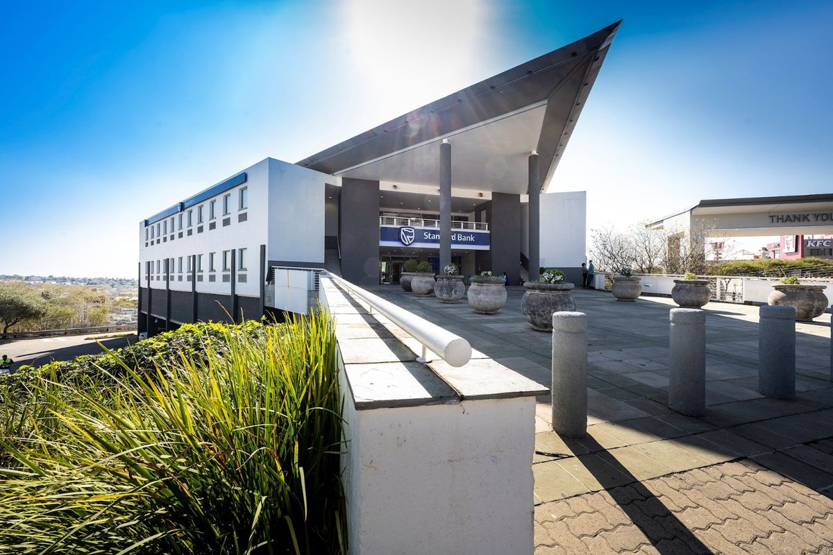Modern commercial building facade featuring a prominent cantilevered roof, concrete support pillars, and a spacious paved entrance terrace.