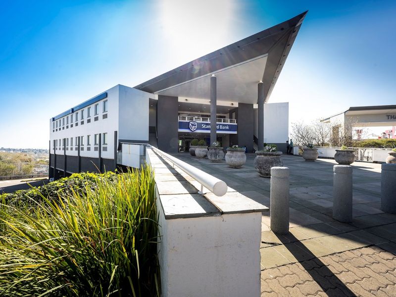 Modern commercial building facade featuring a prominent cantilevered roof, concrete support pillars, and a spacious paved entrance terrace.