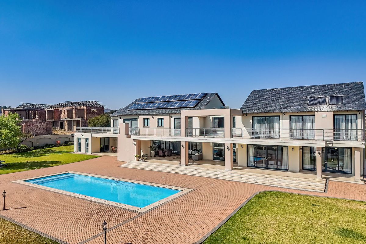 Spacious rear elevation of a modern two-story house featuring a large swimming pool, expansive paved patio, upper-level balcony, and integrated solar panels.