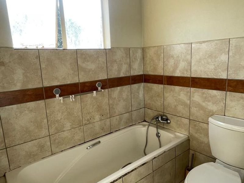 A dated bathroom featuring a standard bathtub, toilet, and neutral-toned wall and floor tiles, requiring cosmetic updates to improve its overall condition.