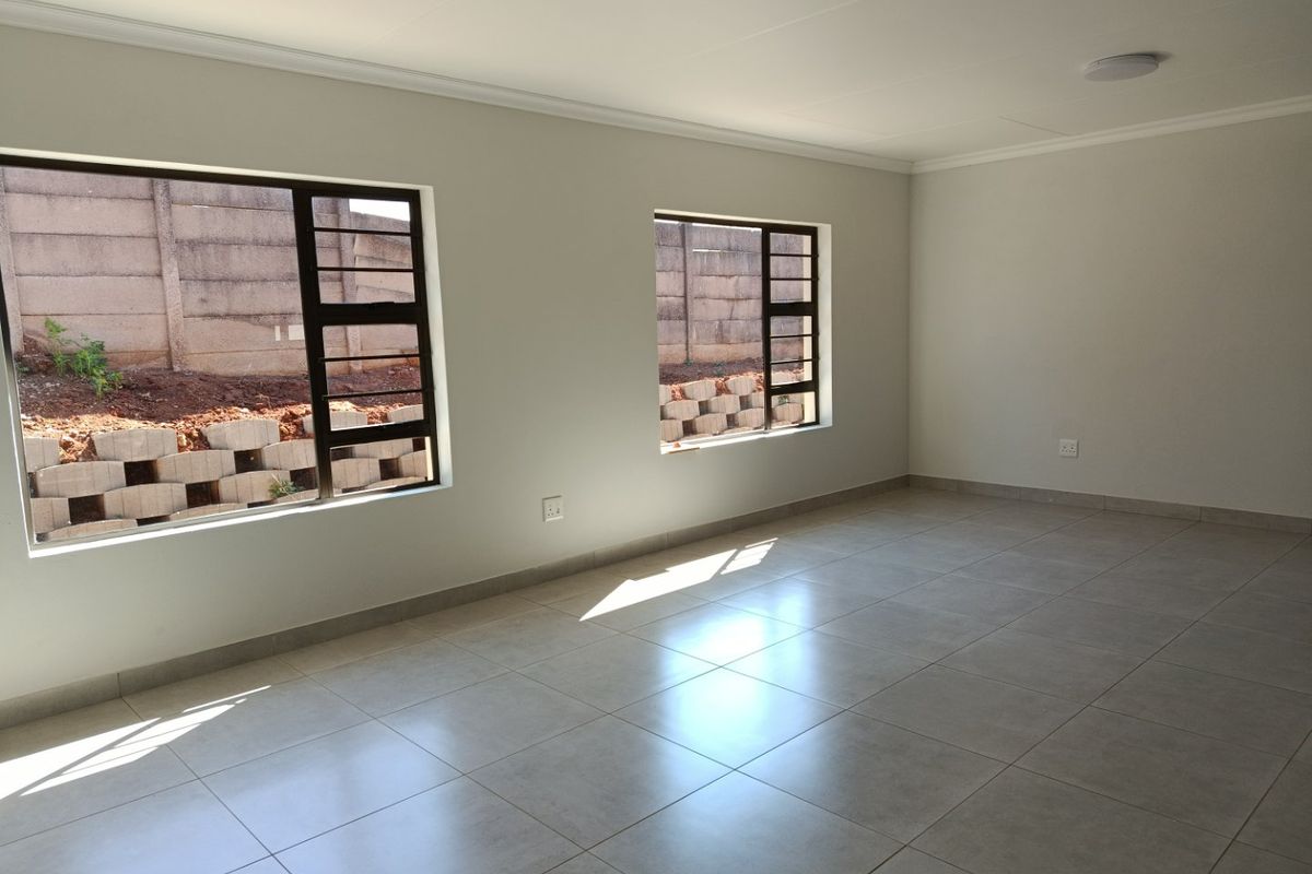 A bright, empty living area featuring neutral tiled flooring, clean white walls, and modern aluminium-framed windows overlooking the rear yard.