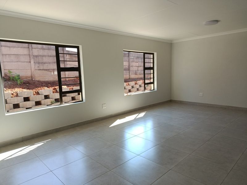 A bright, empty living area featuring neutral tiled flooring, clean white walls, and modern aluminium-framed windows overlooking the rear yard.