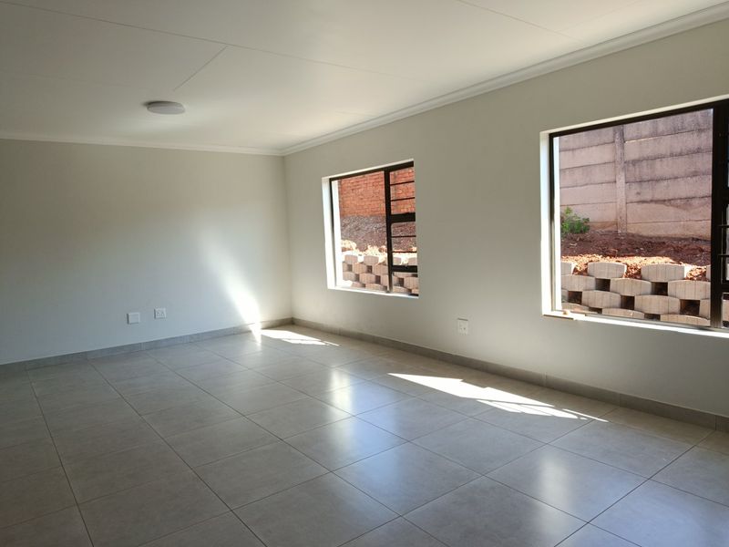 A bright, empty living room featuring neutral tiled flooring, clean white walls, and large windows with views of the rear retaining wall.
