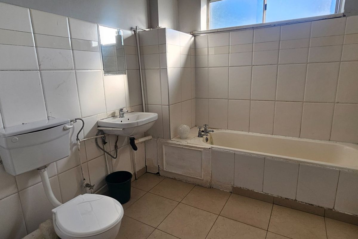 A dated bathroom featuring a built-in bathtub, pedestal basin, and flush toilet, finished with full-height ceramic wall tiling and floor tiles.