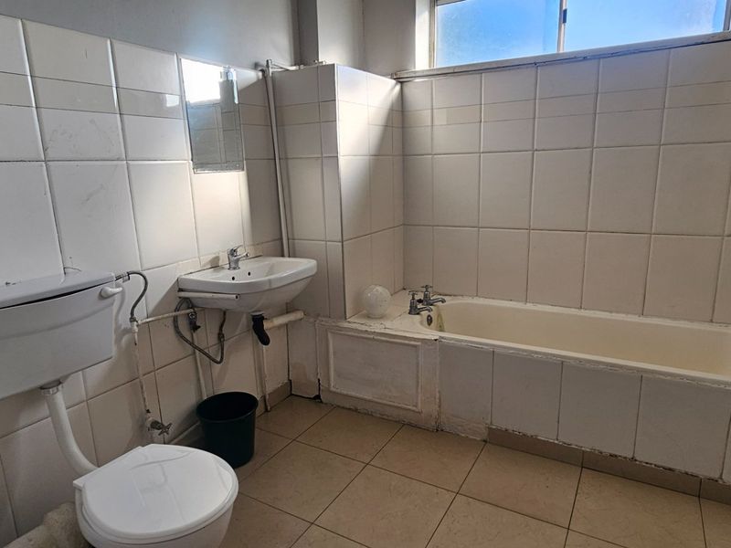 A dated bathroom featuring a built-in bathtub, pedestal basin, and flush toilet, finished with full-height ceramic wall tiling and floor tiles.