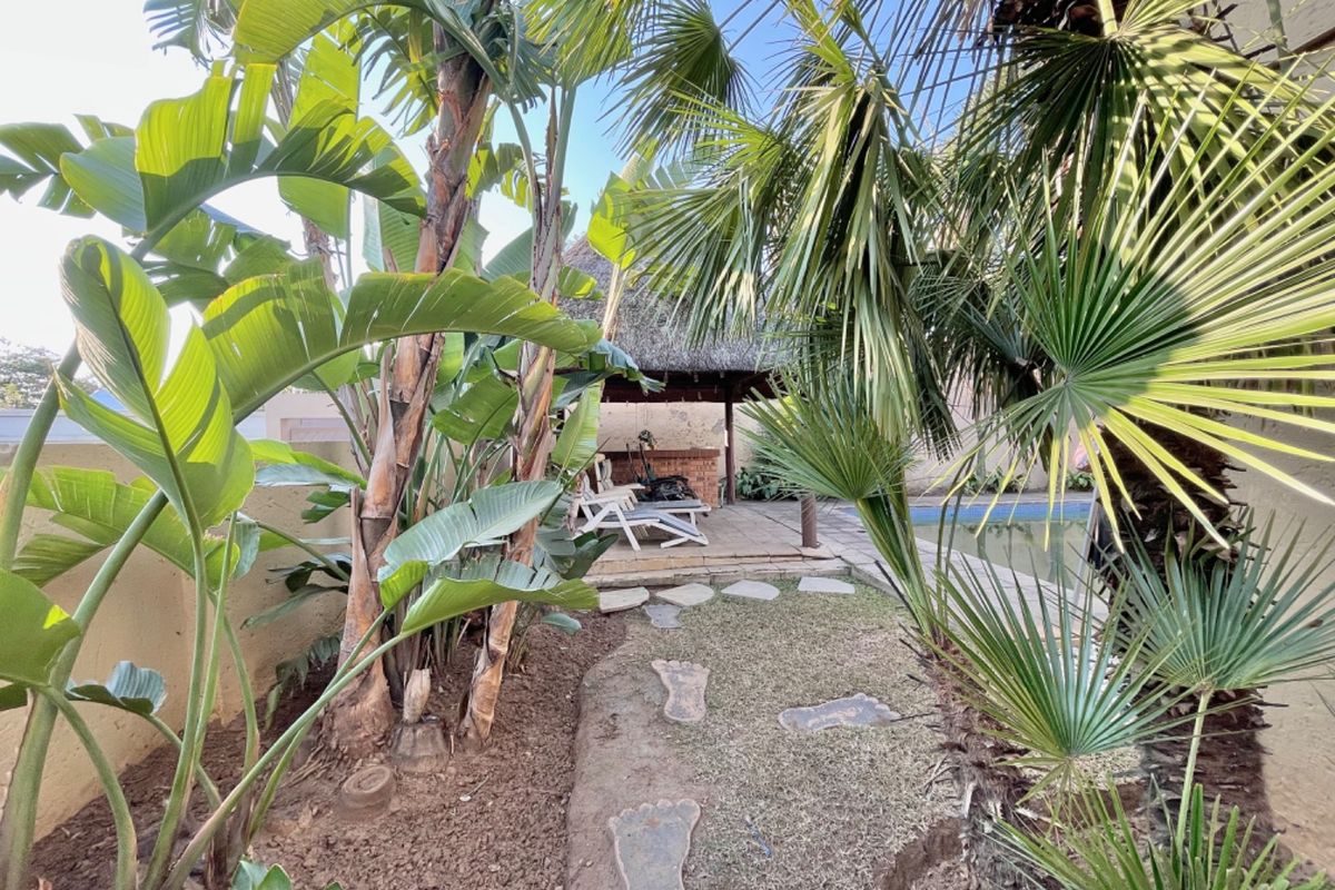 Outdoor pool area featuring a thatched lapa with a built-in brick braai, paved patio space, and established tropical garden landscaping.