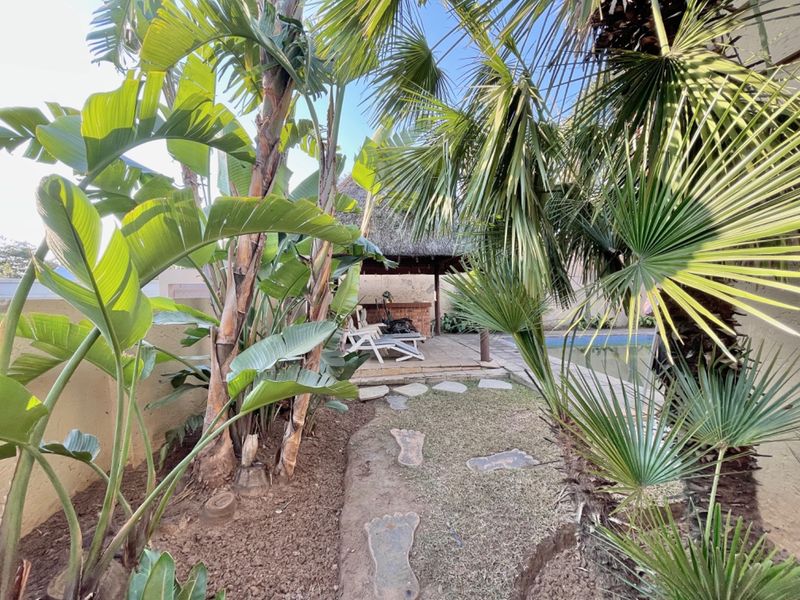 Outdoor pool area featuring a thatched lapa with a built-in brick braai, paved patio space, and established tropical garden landscaping.