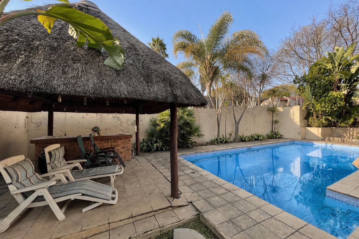 Outdoor pool area featuring a thatched lapa with a built-in brick bar, paved decking, and a swimming pool, set against a perimeter wall.