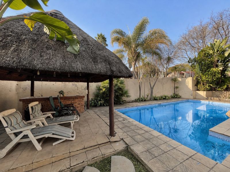 Outdoor pool area featuring a thatched lapa with a built-in brick bar, paved decking, and a swimming pool, set against a perimeter wall.