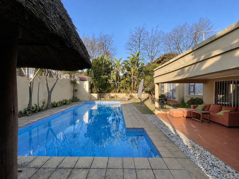 Outdoor pool area featuring a swimming pool, covered patio with tiled flooring, and a thatched lapa structure in a private garden setting.