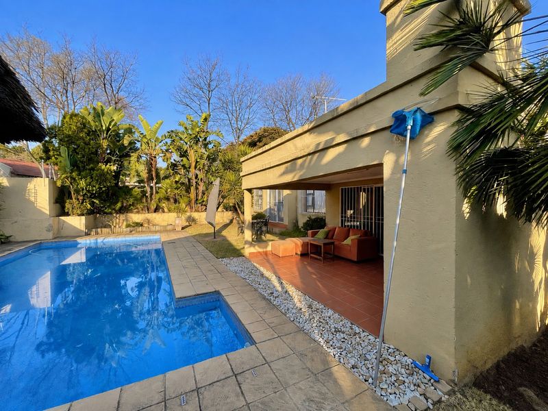 Outdoor entertainment area featuring a swimming pool, tiled patio with a built-in fireplace, and established garden surroundings.