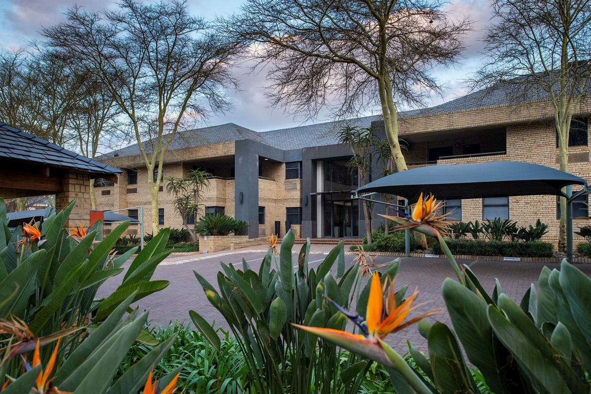 A professional exterior view of a commercial office building featuring a brick facade, paved driveway, and established landscaped gardens with shade sail parking.