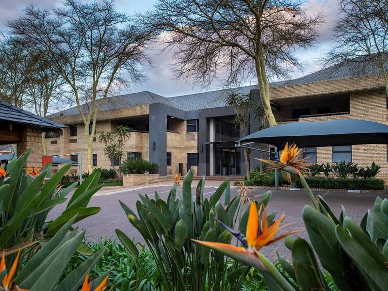 A professional exterior view of a commercial office building featuring a brick facade, paved driveway, and established landscaped gardens with shade sail parking.