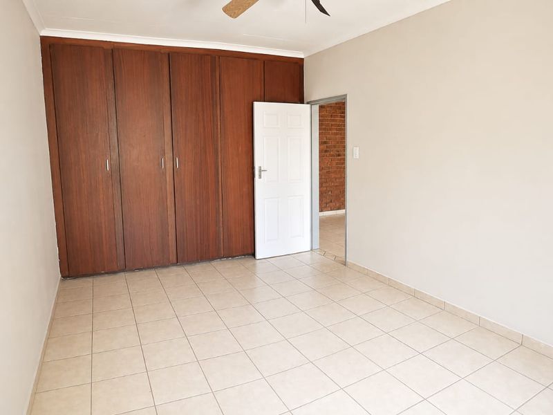 A spacious, empty bedroom featuring neutral tiled flooring, built-in wooden cupboards, and a ceiling fan, offering a clean and versatile living space.