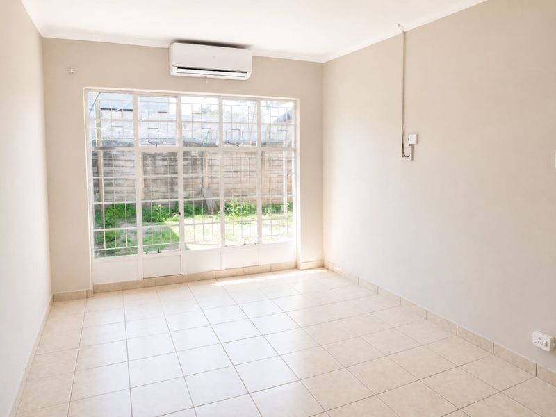 A bright, empty living room featuring tiled flooring, a split-system air conditioner, and a large window with security bars overlooking the rear garden area.