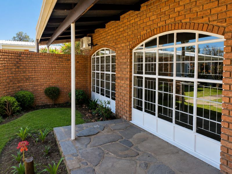 A covered patio area featuring stone paving, exposed brick walls, and large arched windows with security bars overlooking a small garden.