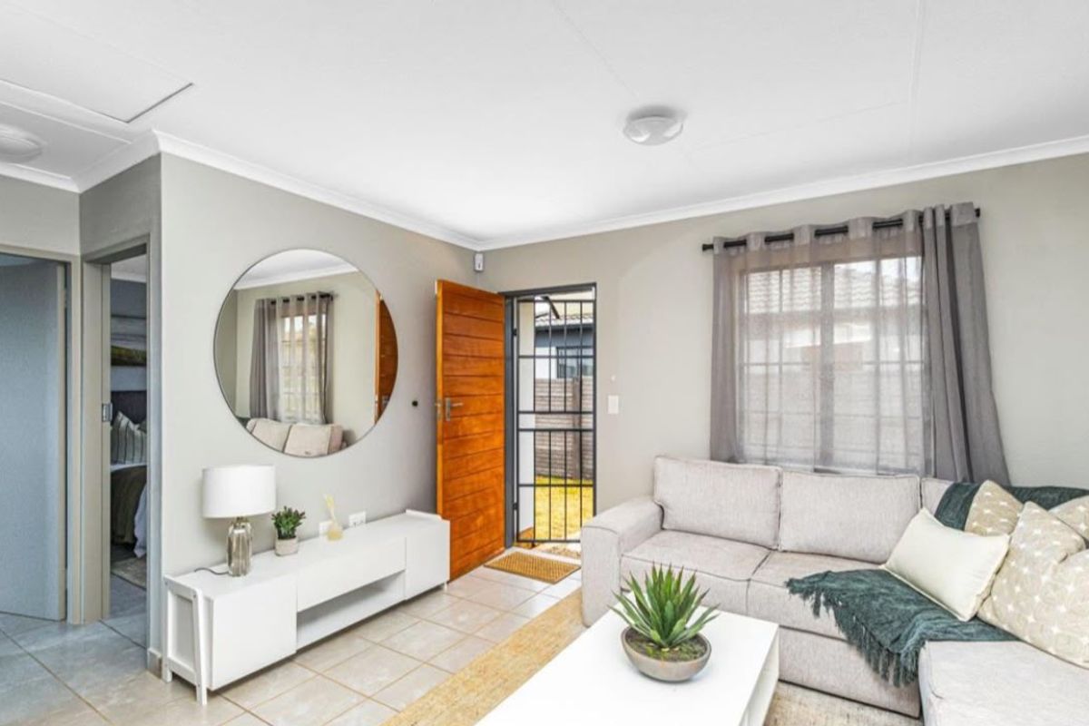 A bright and well-maintained living room featuring tiled flooring, a timber entrance door with a security gate, and neutral wall finishes.