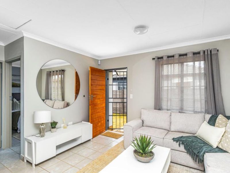 A bright and well-maintained living room featuring tiled flooring, a timber entrance door with a security gate, and neutral wall finishes.