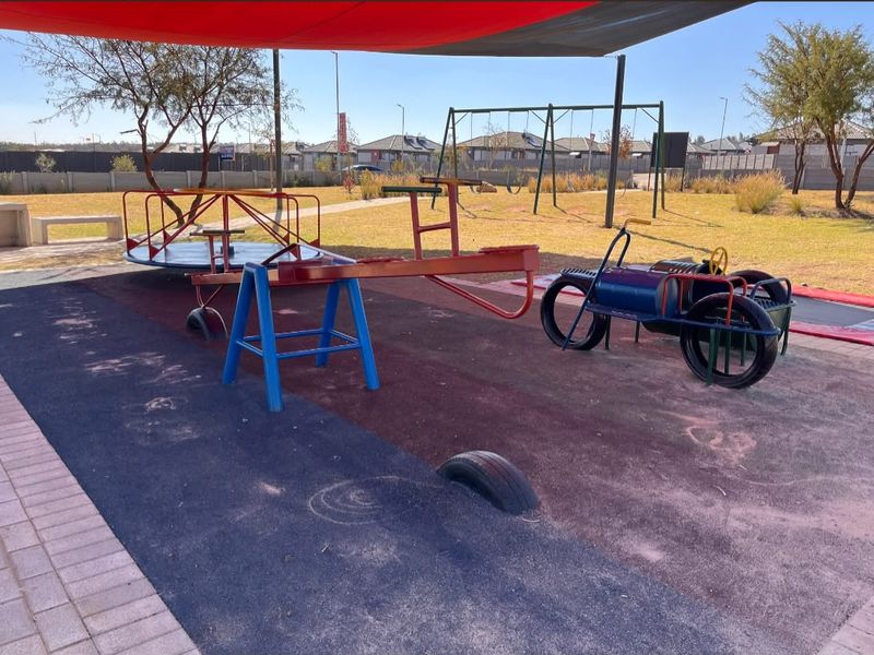 Outdoor communal playground area featuring a shade sail, rubberized flooring, and various fixed play equipment including a merry-go-round and swings.