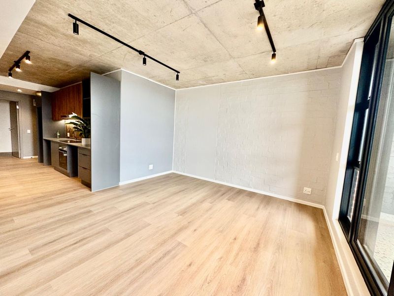 Modern open-plan living area featuring an exposed concrete ceiling, track lighting, laminate flooring, and a sleek kitchen with integrated appliances.
