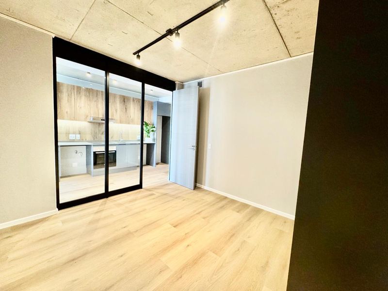 Modern open-plan apartment featuring an exposed concrete ceiling, light wood-look flooring, and a sleek kitchen separated by a stylish black-framed glass sliding partition.