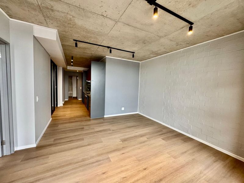 Modern open-plan living area featuring an exposed concrete ceiling, track lighting, laminate flooring, and a stylish painted brick feature wall.