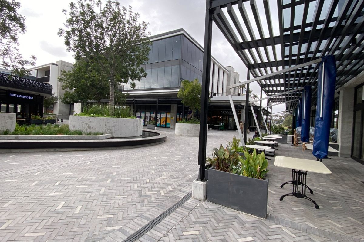 A modern outdoor courtyard featuring herringbone paving, concrete planters, and a metal pergola structure, set against a contemporary glass-fronted commercial building.