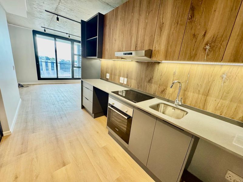 Modern open-plan kitchen featuring sleek cabinetry, stone countertops, integrated appliances, and an exposed concrete ceiling, flowing into a bright living area with balcony access.