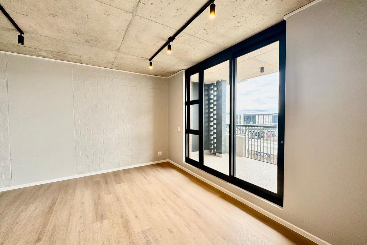 Modern, empty living area featuring an exposed concrete ceiling, track lighting, laminate flooring, and a large sliding glass door leading to a balcony.