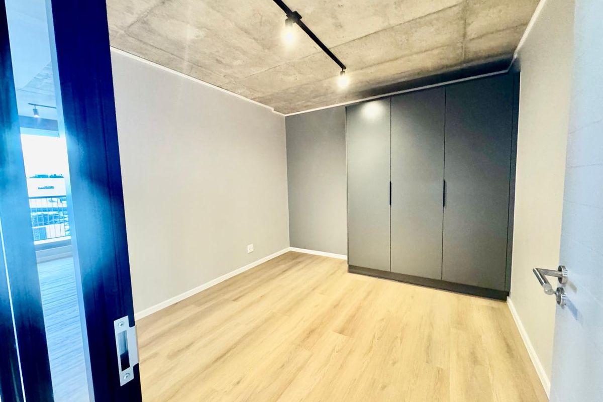 A modern, empty bedroom featuring light laminate flooring, a sleek dark grey built-in wardrobe, and an industrial-style exposed concrete ceiling with track lighting.