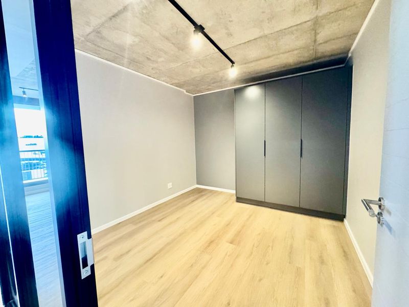 A modern, empty bedroom featuring light laminate flooring, a sleek dark grey built-in wardrobe, and an industrial-style exposed concrete ceiling with track lighting.