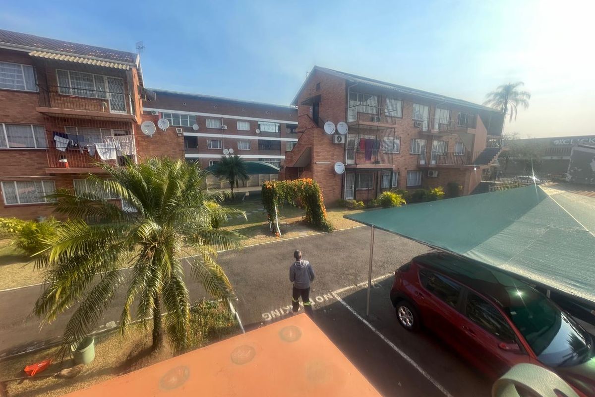View of the apartment complex exterior featuring brick facades, private balconies, a paved parking area with carports, and communal garden space.