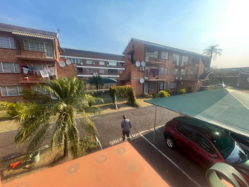 View of the apartment complex exterior featuring brick facades, private balconies, a paved parking area with carports, and communal garden space.