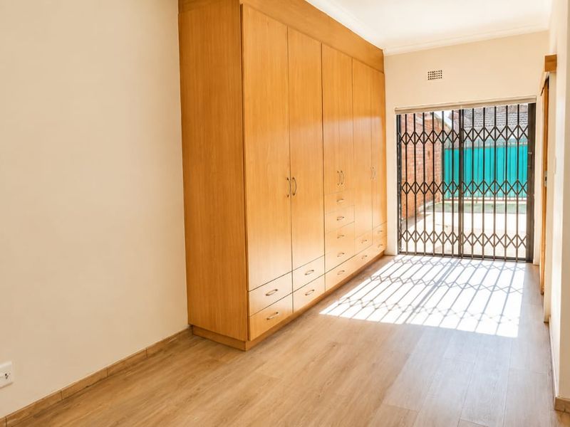 A bright, empty bedroom featuring light wood-look laminate flooring and extensive built-in wooden cabinetry, with direct access to an outdoor area via a security gate.