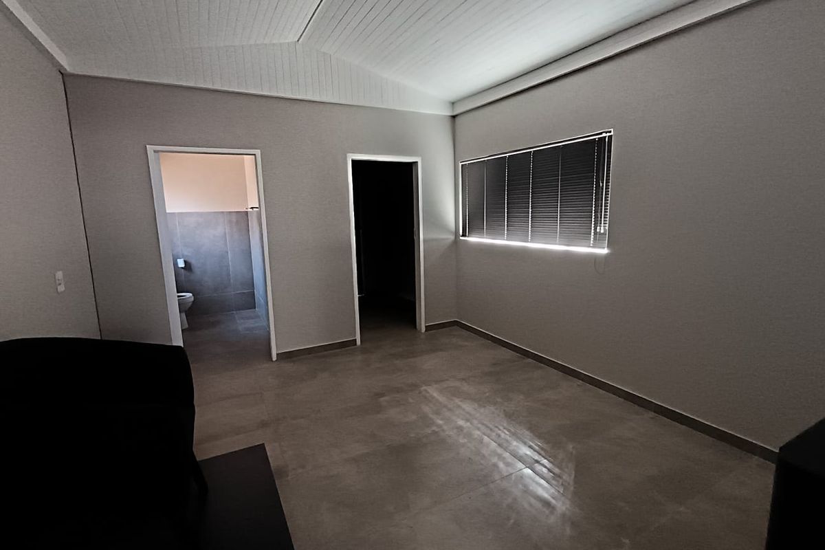 A clean, minimalist bedroom featuring tiled flooring, a vaulted ceiling, and neutral walls, with an adjoining bathroom visible through the doorway.