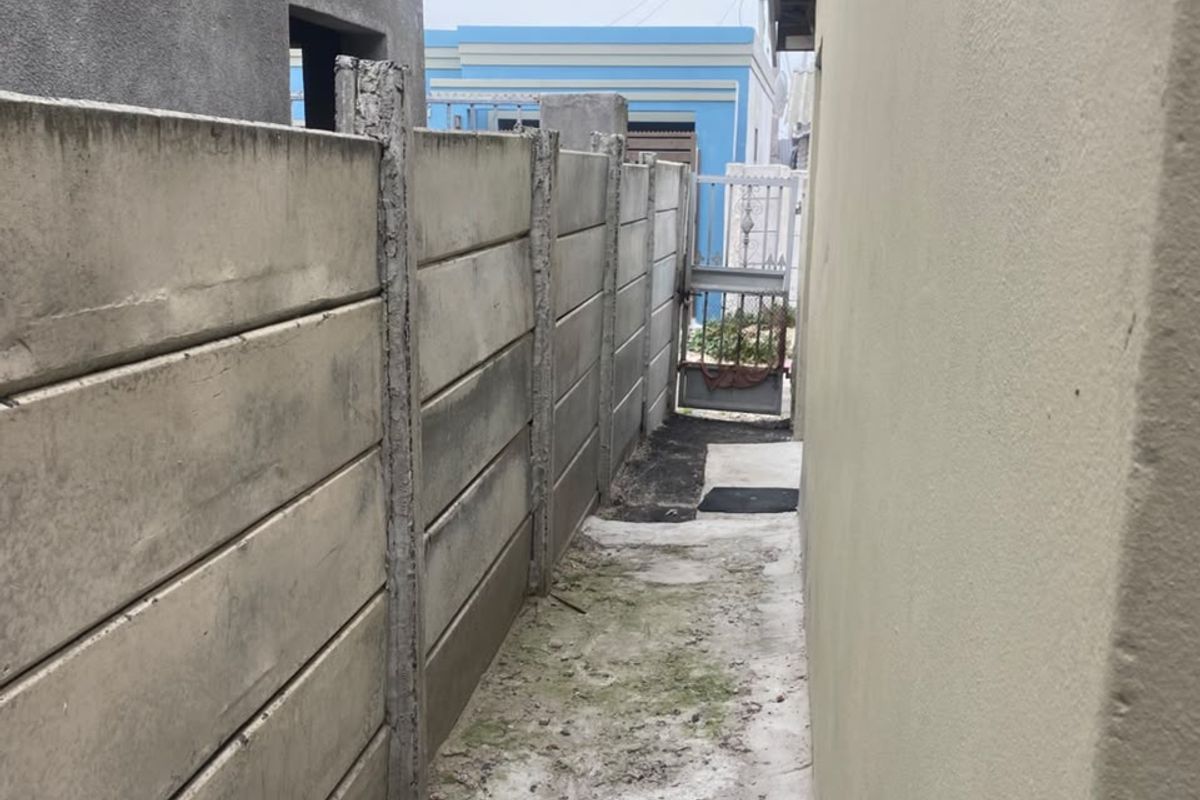 A narrow side passage featuring a precast concrete boundary wall, metal security gate, and weathered concrete pathway.