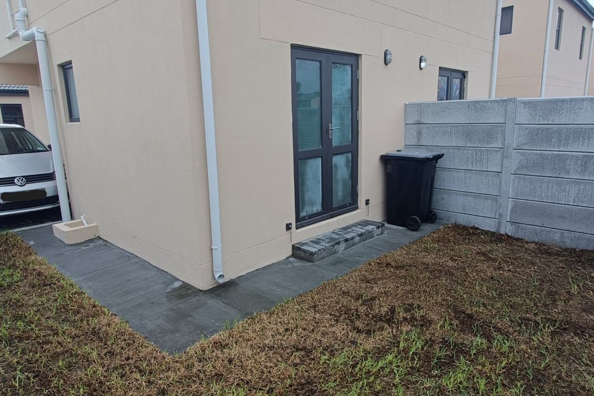 Rear exterior view of the townhouse featuring a paved walkway, double glass door access, and a precast concrete boundary wall with an unfinished garden area.