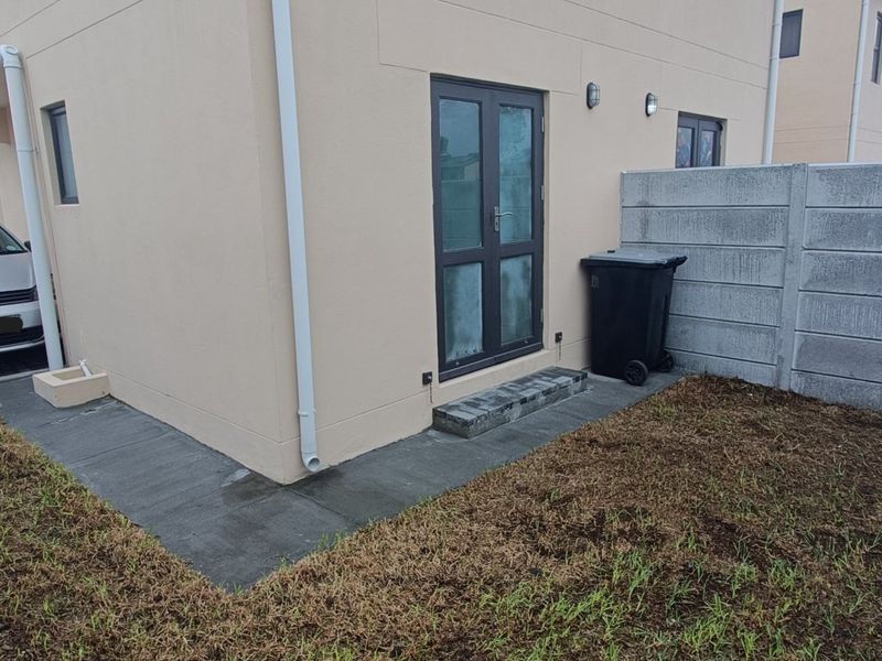Rear exterior view of the townhouse featuring a paved walkway, double glass door access, and a precast concrete boundary wall with an unfinished garden area.