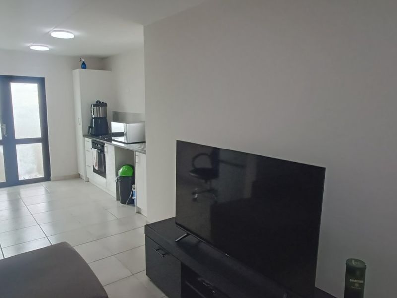 Open plan living area featuring tiled flooring and a compact kitchen with built-in cabinetry, oven, and hob, leading to a glass door entrance.