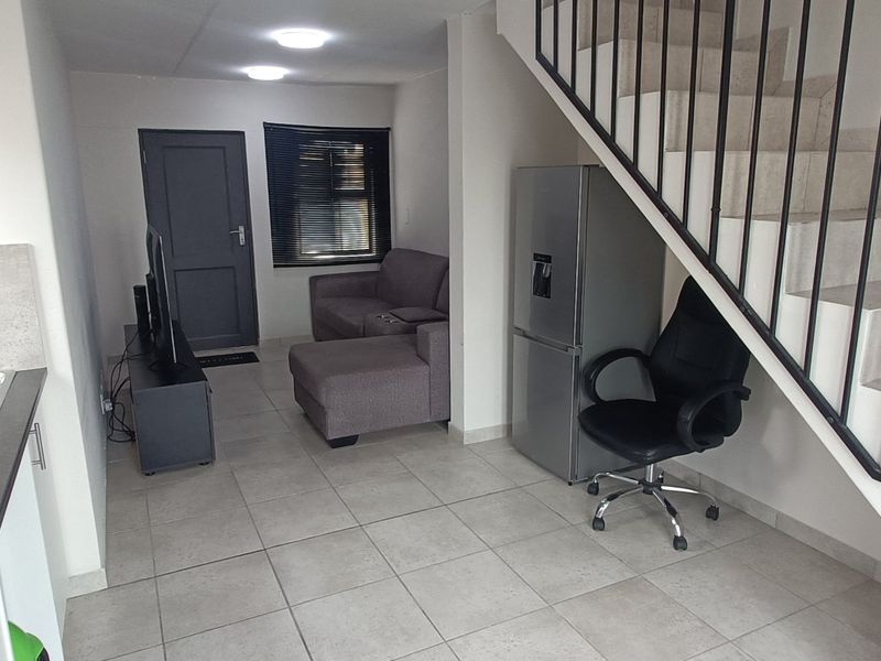 Open plan living area featuring tiled flooring, a staircase with a metal railing, and a compact kitchen section with a sink and countertop.
