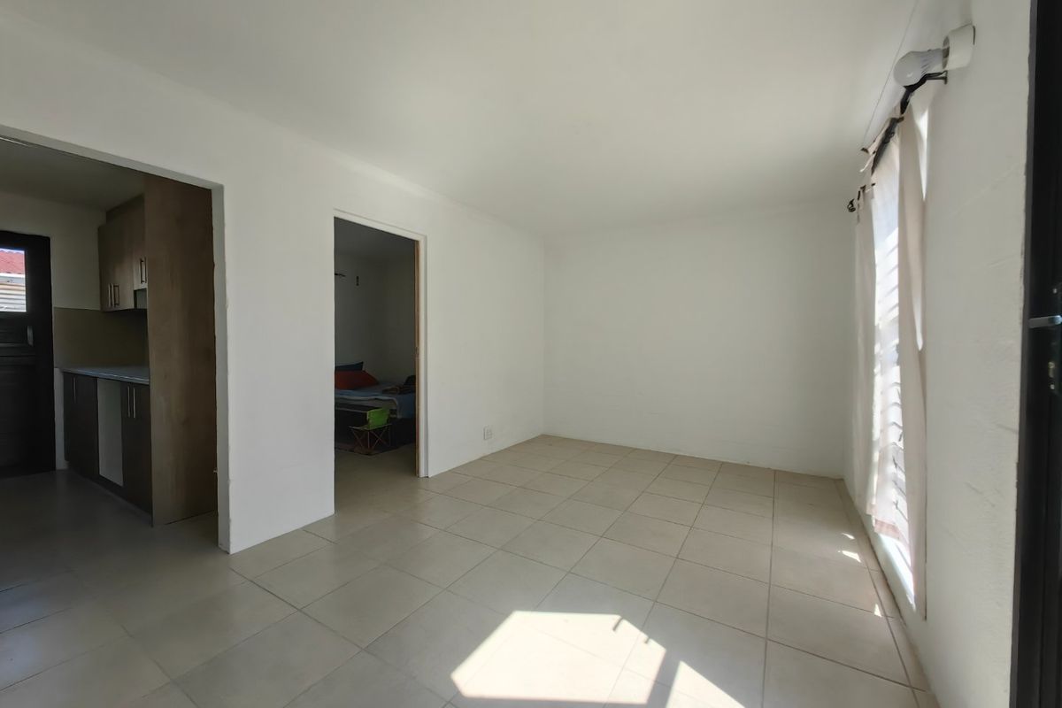 A bright, empty living area featuring neutral tiled flooring and natural light from a side window, with an adjacent kitchen visible.