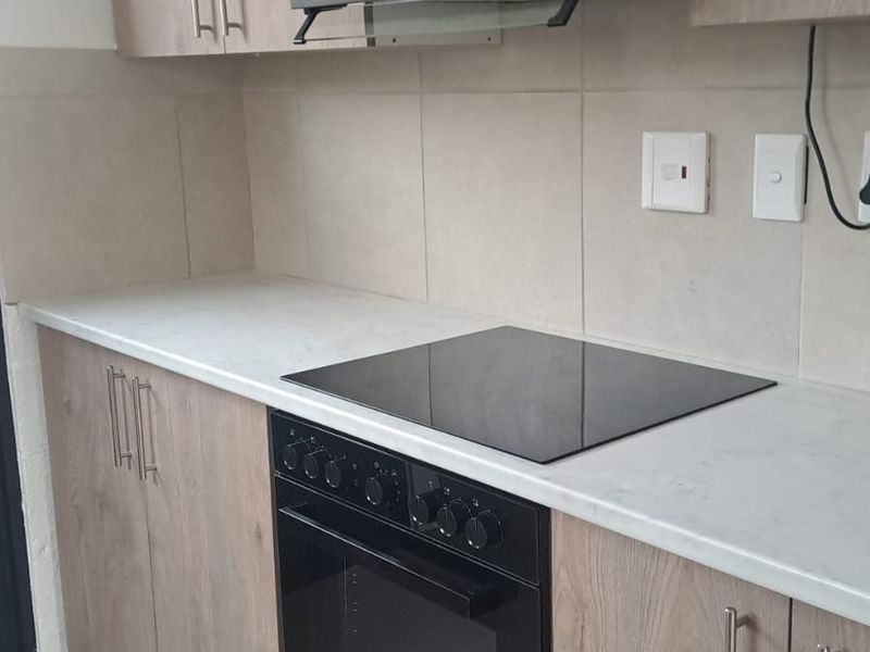 A modern kitchen featuring wood-finish cabinetry, stone-look countertops, a built-in oven, electric hob, and a sleek extractor fan with tiled backsplash.