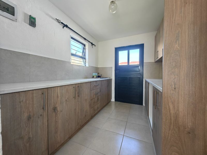 A clean and functional scullery featuring modern wood-look cabinetry, stone-style countertops, tiled splashbacks, and direct access to the exterior via a secure door.