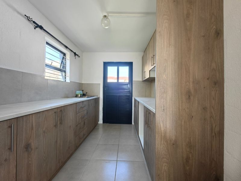 A clean, galley-style kitchen featuring modern wood-look cabinetry, neutral tiled flooring, and a functional layout with direct exterior access.