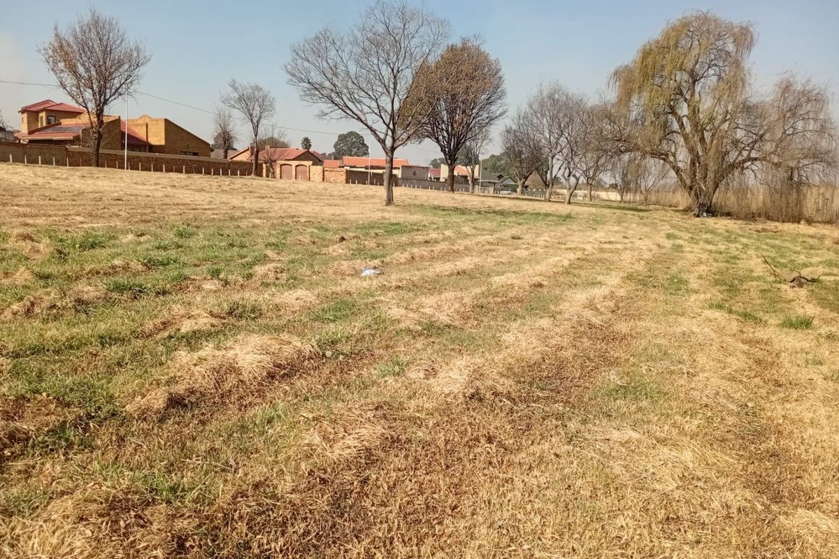 A view of a vacant residential plot featuring open grassland and mature trees, with neighboring residential properties visible in the background.