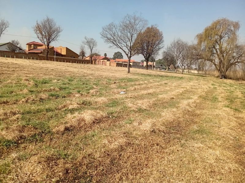 A view of a vacant residential plot featuring open grassland and mature trees, with neighboring residential properties visible in the background.
