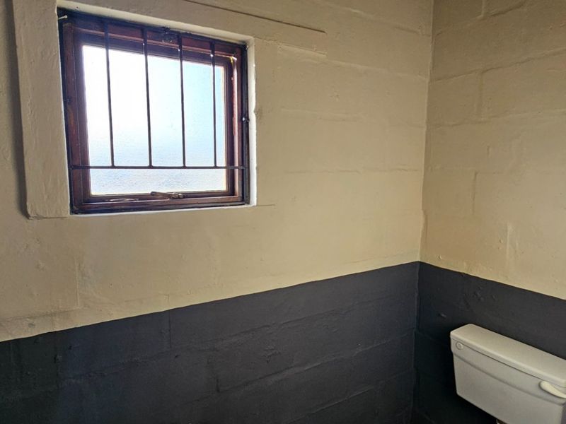 A small, functional bathroom featuring exposed masonry walls, a toilet, and a window with security bars.