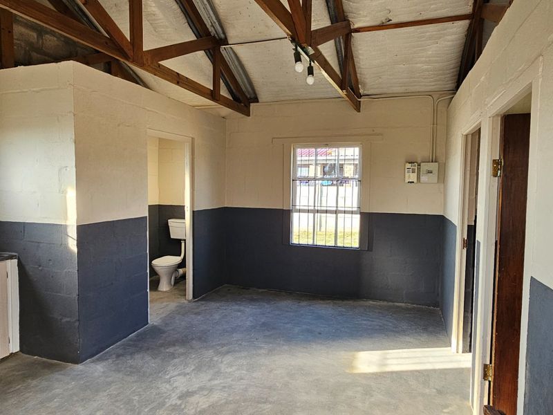 Open-plan living area featuring exposed timber roof trusses, concrete flooring, a built-in sink unit, and an adjoining private toilet facility.